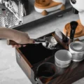 Hand hält den Siebträger über eine dunkle Organizer-Box aus Holz mit Puck Screen, Bürsten und zwei Distributoren
