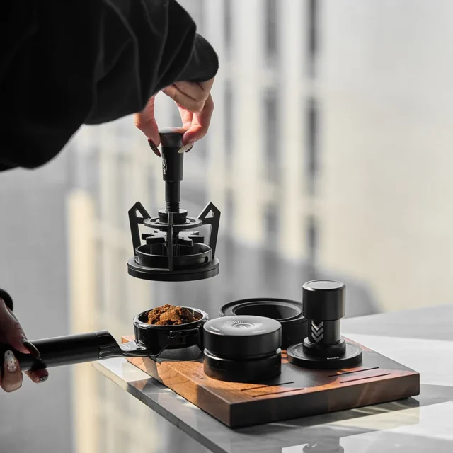 Hand hebt das V3 WDT-Tool über Tamper und Distributor des Sets und einen Siebträger mit klumpigem Kaffeemehl