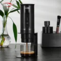 Schwarze tragbare IKAPE Espressomaschine im Ständer gießt in ein Glas, auf einem dunklen Holztisch neben einer Pflanze