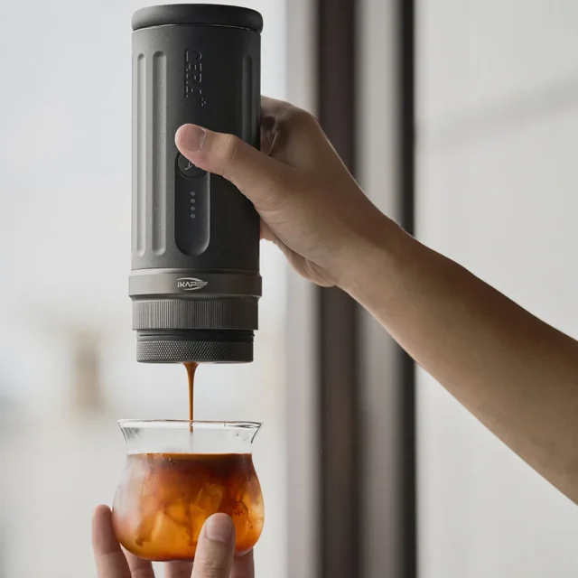 IKAPE Kapo K2 in einer Hand am Fenster, Espresso läuft aus dem Brühkopf in ein Glas mit Eis