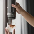 K2 in einer Hand, der Daumen auf dem Knopf, Espresso tropft in ein Glas mit Milch und Eis