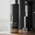 K2 aufrecht auf einem Holztisch, die graue Tasse unten aufgesetzt, dahinter unscharf eine Mühle