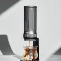 Graue K2 Pro im schwarzen Ständer brüht Espresso auf Eis im Glas, in hartem Sonnenlicht vor einer weißen Wand