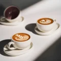 Zwei Latte-Tassen mit Rosetta in tiefem Sonnenlicht, dahinter eine dritte, innen dunkel glasiert, gekippt auf der Untertasse