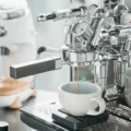 Latte-Tasse auf schwarzer Waage unter einer verchromten Espressomaschine mit Manometern, Espresso läuft aus dem Siebträger