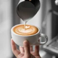 Milch fließt aus einem Milchkännchen aus Stahl in eine Latte-Tasse in einer Hand und zeichnet ein Herz