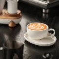 Latte-Tasse mit Schwanenmuster auf ihrer ovalen Untertasse, Milchkännchen und weiße Mühle unscharf