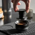 Hand hält den Tamper über den Trichter, darunter Kaffeemehl im Sieb
