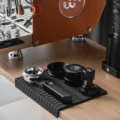 Polaris Siebträger auf einer schwarzen Tampermatte mit Trichter, Distributor und Tamper, dahinter Maschine und Mühle