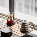 Schwarze IKAPE Keramikkochplatte auf einem Holzlattentisch neben Pour-over-Glaskaraffe und Schwanenhalskanne aus Stahl