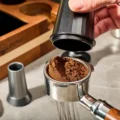 Hand hebt den Dosierbecher von einem Siebträger, im Sieb bleibt eine gehäufte Dosis Kaffeemehl