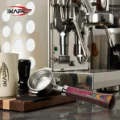 Rainbow Bottomless-Siebträger auf einer Arbeitsfläche vor einer Espressomaschine, links ein Kännchen und ein Tamper