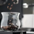 Kaffeebohnen fallen in ein leeres Sense-Glas auf einem kleinen schwarzen IKAPE Sockel, dahinter unscharf eine Maschine