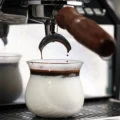 Espresso tropft aus den Ausläufen eines Siebträgers mit Holzgriff in ein Sense-Glas mit Milch