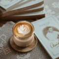 Sense-Glas mit Latte und Tulpenmuster auf rundem Holzuntersetzer, auf einer Spitzendecke neben einem Bücherstapel