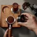 Draufsicht: Siebträger mit Kaffeemehl in der vorderen Aufnahme der Station, eine Hand bringt einen Distributor