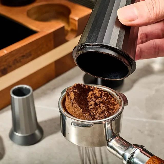 Hand hebt einen schwarzen Dosierbecher von einem Siebträger mit gehäuftem Kaffeemehl, dahinter ein Organizer aus Holz