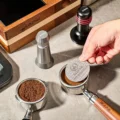 Hand legt das Gewebe-Puck-Screen auf getampten Kaffee in einem Siebträger, daneben ein zweiter mit Kaffeemehl