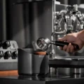 Hand hält einen leeren Siebträger über den V2 Behälter, ein ausgebrauchter Puck auf dem Block, dahinter eine Espressomaschine