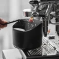 Abklopfbehälter V2 unter einer Brühgruppe fängt Wassertropfen auf, während eine Bürste die Gruppe reinigt