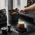 Behälter voller Pucks auf schwarzer Arbeitsfläche, darüber ein Siebträger, vorne ein Glas Espresso auf einer Waage