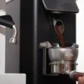 Silberner IKAPE Dosiertrichter auf einem Siebträger in einer schwarzen Mühle, fängt Kaffeemehl auf