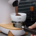 Weißer IKAPE Dosiertrichter auf einem Siebträger unter dem Auswurf einer weißen Mühle, Kaffee fällt hinein