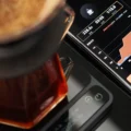 Schwarze IKAPE V2 Pro Waage unter einer Karaffe, daneben ein Handy mit 228,6 g, 03:05 und Verhältnis 1:15,0