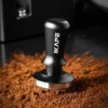 Schwarzer V3 Tamper auf verstreutem, lockerem Kaffeemehl, dahinter eine dunkle Küche