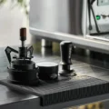 Schwarzer V3 Tamper auf schwarzer Matte mit Wellenmuster neben einem Distributor und einem WDT-Tool im Ständer