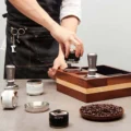 Barista in Jeansschürze setzt einen schwarzen Distributor auf einen Siebträger im Holz-Organizer, vorne silberne Werkzeuge