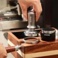 Hand senkt den silbernen V3 auf einen Siebträger mit Kaffee in einem Holz-Organizer, daneben ein schwarzer Distributor