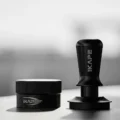 Ganz schwarzer IKAPE V3 Tamper neben einem gerändelten schwarzen Distributor auf heller Fläche