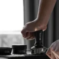 Hand drückt den schwarzen IKAPE V3 Tamper in einen Siebträger auf einer geriffelten Matte, daneben Trichter und Distributor
