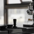 Schwarzer IKAPE V3 Tamper auf einer geriffelten Matte neben Trichter und Distributor, dahinter läuft ein Espresso