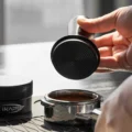 Hand hält den schwarzen IKAPE V3 Tamper mit dem Boden nach unten über ein Siebträgersieb mit Kaffeemehl