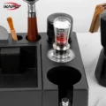 Tamper mit klarem Griff in einer schwarzen Tamperstation, daneben eine Bürste und ein Siebträger mit Holzgriff