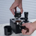 Hand senkt den Distributor auf ein Sieb mit Kaffeemehl in der Box, daneben eine schwarze geschlitzte Hülse