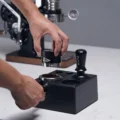 Hand hält den Distributor über einen Siebträger mit Kaffeemehl in der Box, dahinter der Tamper aufrecht