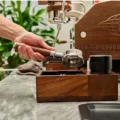 Hand hält einen Siebträger mit Holzgriff über die Nussbaumstation, dahinter Espressomaschine und Pflanzen