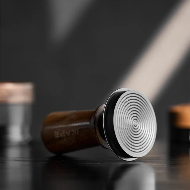 IKAPE Tamper mit Nussbaumgriff liegend, die silberne Basisfläche mit konzentrischen Ringen zur Kamera