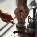 Hand drückt den Tamper mit Nussbaumgriff in einen Siebträger, die andere Hand hält dessen Griff, daneben weitere Werkzeuge