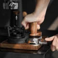 Hand tampt mit dem V3 in einen Siebträger auf einer Nussbaumstation, IKAPE auf dem Griff graviert, links ein Distributor