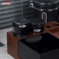 Schwarzer V4 Tamper und schwarzer Distributor nebeneinander auf einer hölzernen Abklopfstation, dahinter eine Kaffeedose