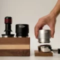 Hand senkt einen silbernen V4 auf einen Siebträger, ein schwarzer V4 und ein anderer Tamper auf einem Holzblock
