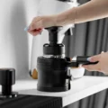 V4 Ständer auf einer weißen Arbeitsfläche hält einen Siebträger, eine Hand tampt, dahinter eine Espressomaschine