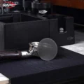 Silbernes V4 Screen an einem Siebträger auf schwarzer gerippter Matte, dahinter eine schwarze Abklopfstation