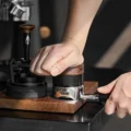 Handfläche drückt auf den Nussbaumkopf des V4, der einen Siebträger auf einem Holztablett tampt