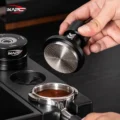 Hand kippt den V6 Mini und zeigt die gerillte Stahlfläche über einem Siebträger mit getamptem Kaffee in einer Station