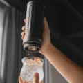Hand hält ein schwarzes tragbares IKAPE Brühgerät aufrecht über ein Glas mit Eis, Espresso tropft heraus